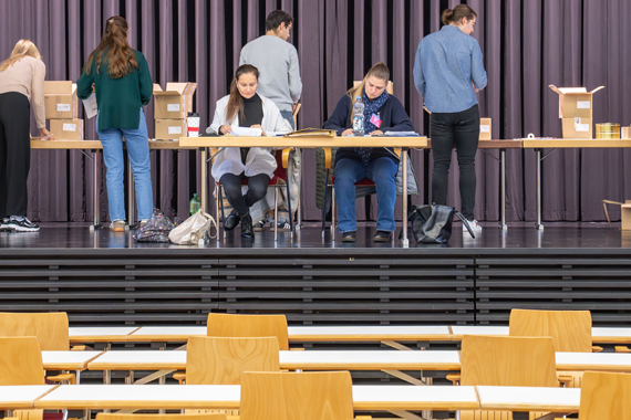 Auf einer Bühne in einer Halle zwei Personen als Testleitungen an Tischen und überwachen den Raum.